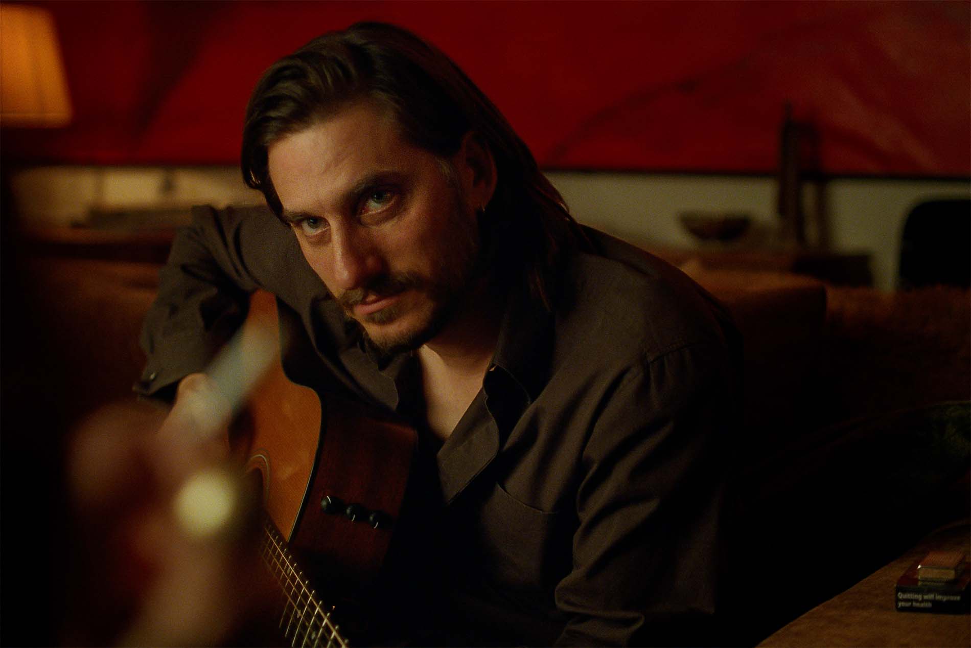 Luca Marinelli in una scena di The Echo Chamber, con una chitarra in una stanza dalle luci rosse