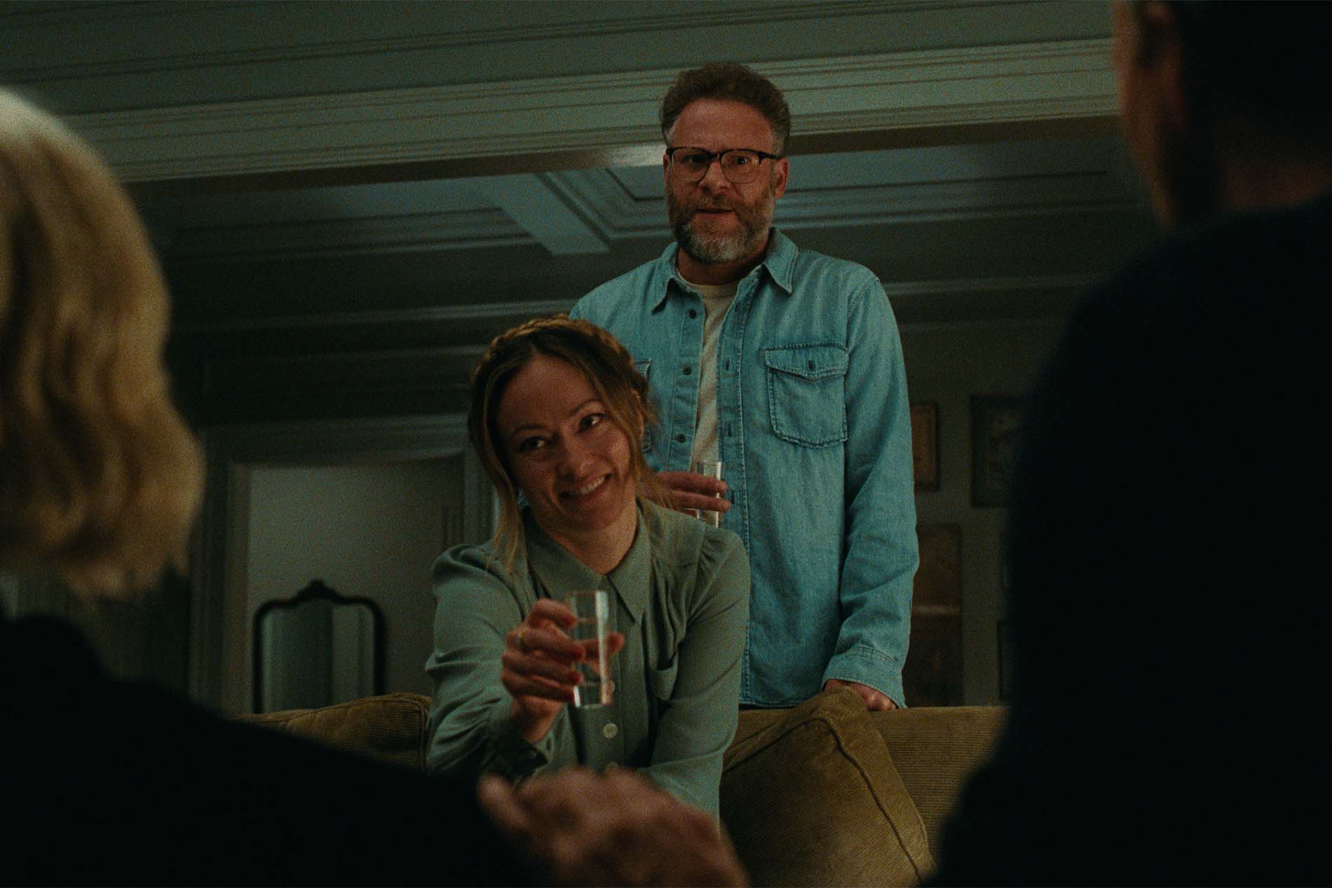 Scena di The Invite - Il Piacere è Tutto Nostro: Olivia Wilde sorride con un bicchiere in mano, mentre Seth Rogen le sta dietro in salotto.