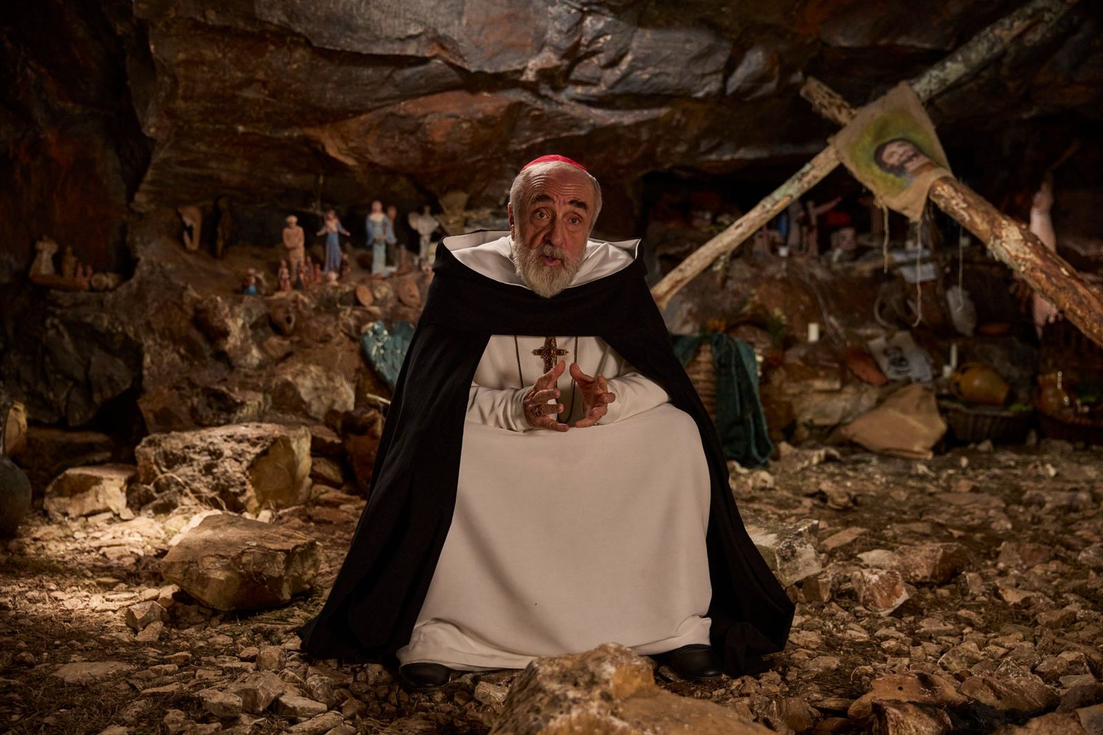 Il cardinale Maculani, in abito bianco e mantello nero, siede in una grotta piena di statuine votive e parla gesticolando.