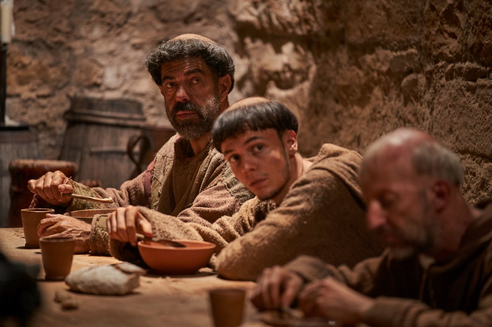 Frate Leopoldo a tavola con altri frati in un refettorio di pietra, tra ciotole di terracotta e pane.