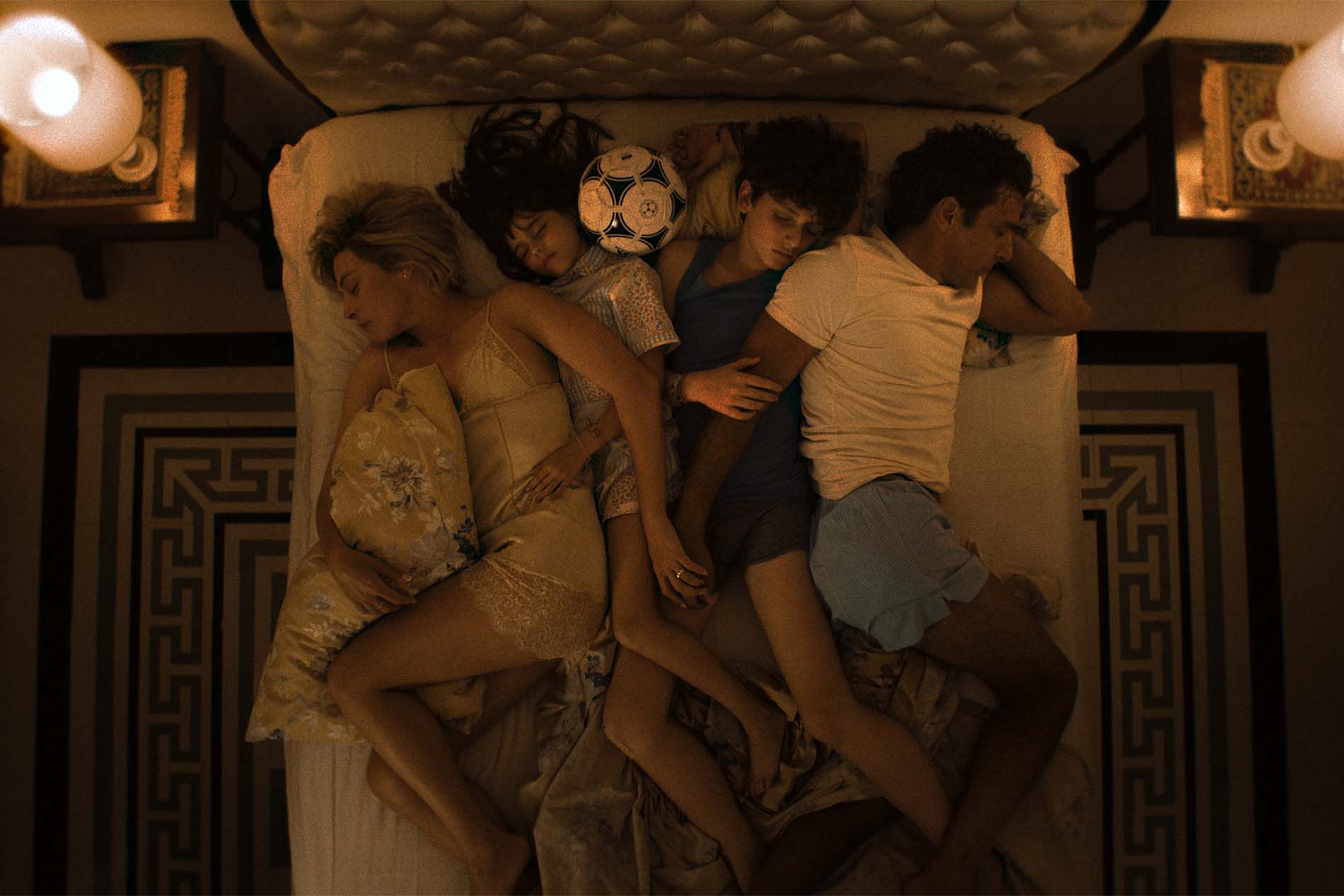 Vista dall’alto: una famiglia addormentata tutta insieme in un grande letto, tra cuscini, lenzuola e un pallone da calcio.