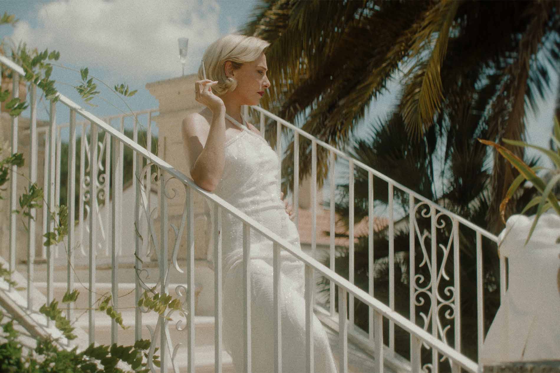 Una donna in abito bianco sulla scalinata di una villa bianca, tra le palme, con una mano all’orecchino.